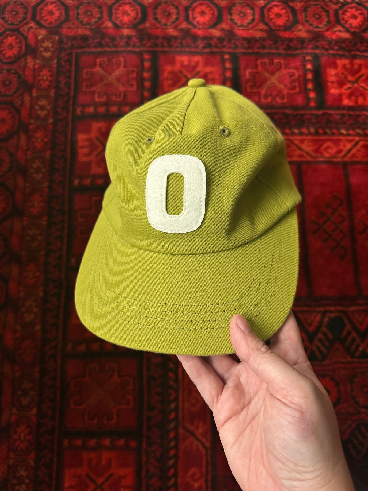On Our Time's "The Hat" baseball cap, in the yellow-green colour called "Olive Sun." The photo is showing the front of the hat and the backdrop is a bright red Persian rug.