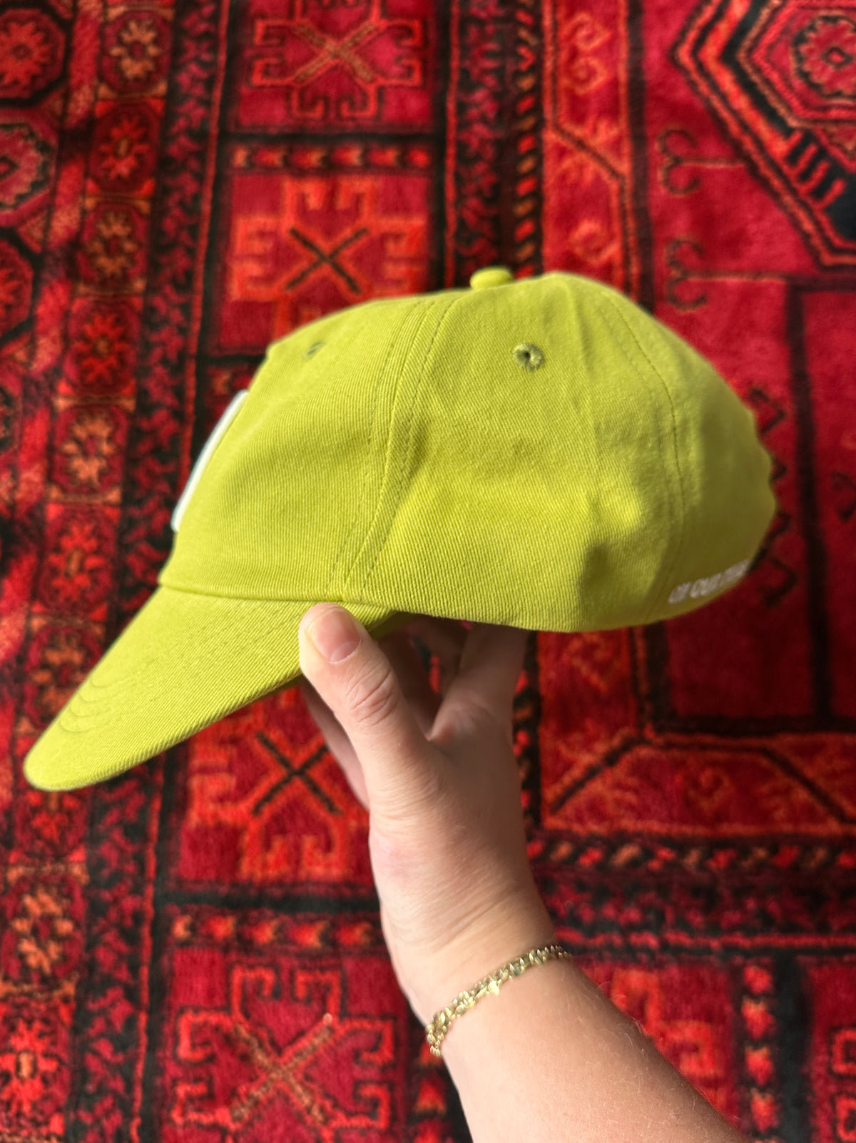 On Our Time's "The Hat" baseball cap, in the yellow-green colour called "Olive Sun." The photo is showing the side of the hat and the backdrop is a bright red Persian rug.