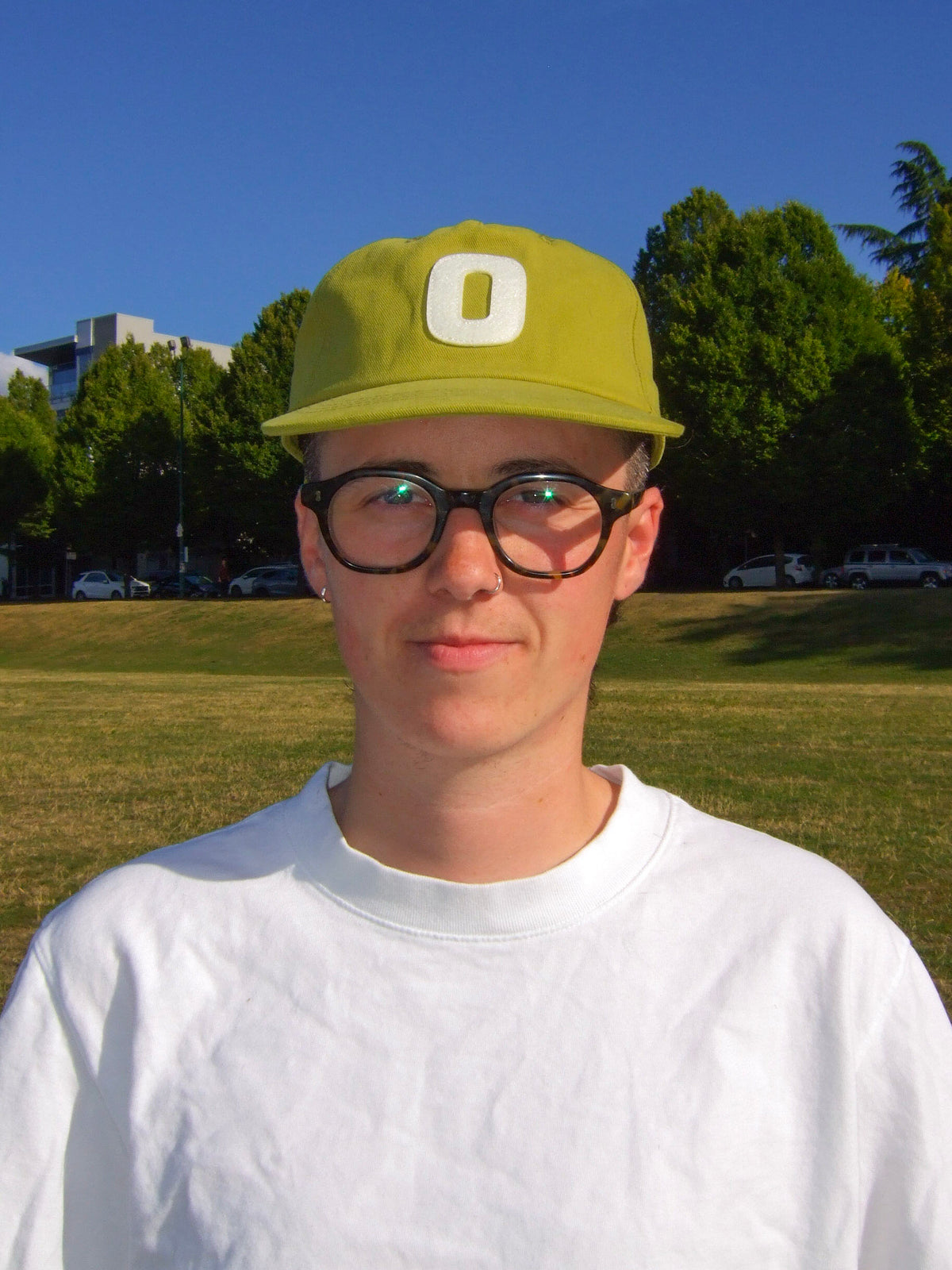 A masculine presenting non-binary person wearing On Our Time's "The Hat" baseball cap, in the yellow-green colour called "Olive Sun." They are wearing dark rimmed glasses and a white t-shirt. The photo is taken of their front. The background is a grass park, trees, and a blue sky. The colours are very warm and saturated. 