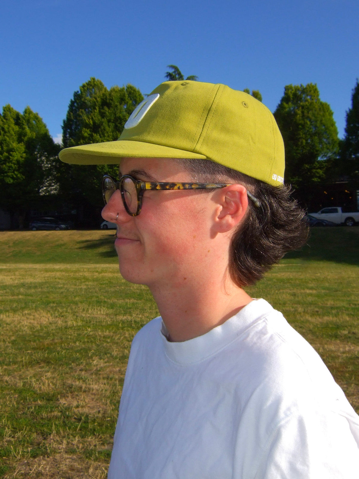 A masculine presenting non-binary person wearing On Our Time's "The Hat" baseball cap, in the yellow-green colour called "Olive Sun." They are wearing dark rimmed glasses and a white t-shirt. The photo is taken of their side profile. The background is a grass park, trees, and a blue sky. The colours are very warm and saturated. 