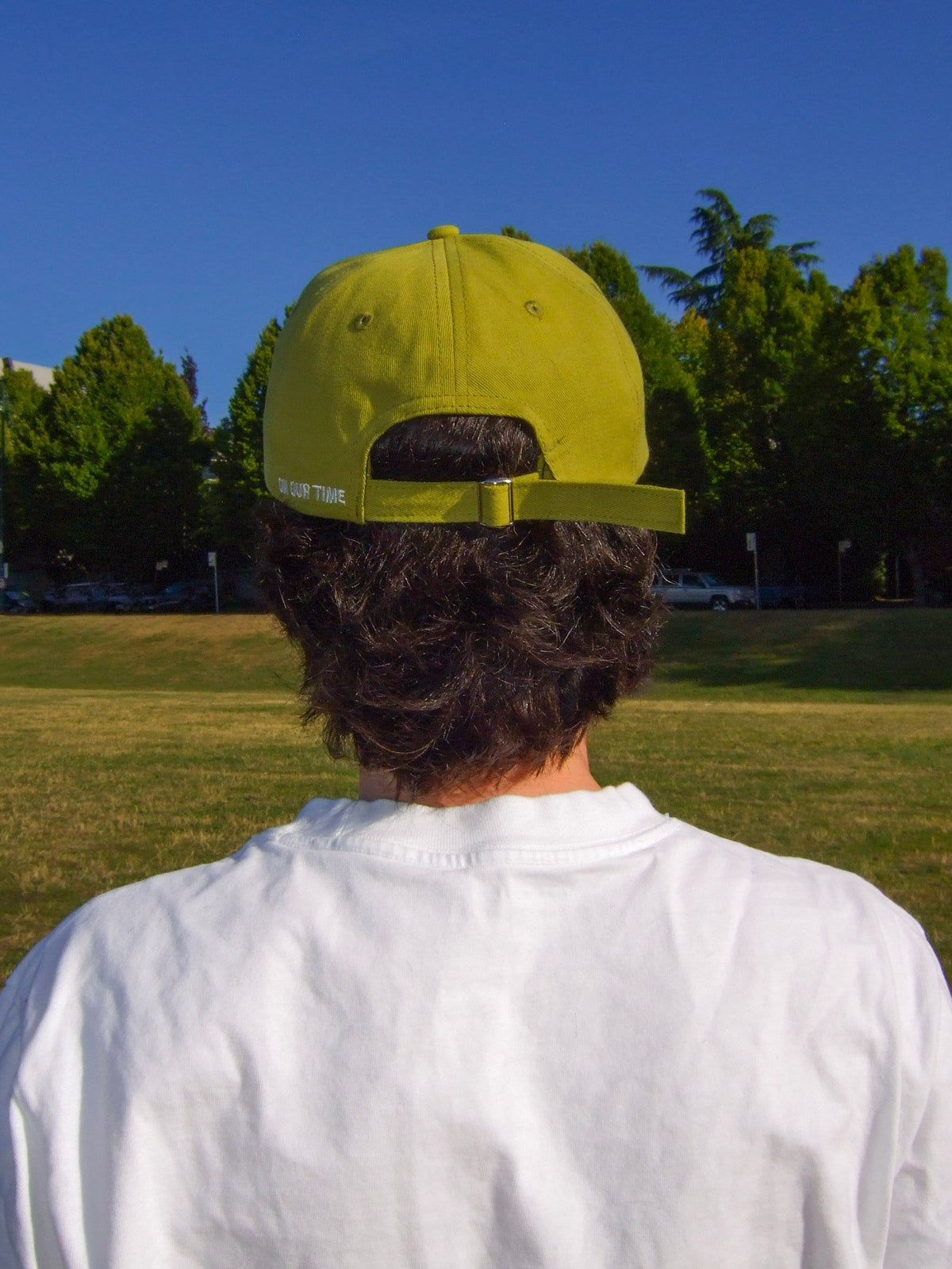 A masculine presenting non-binary person wearing On Our Time's "The Hat" baseball cap, in the yellow-green colour called "Olive Sun." They are wearing dark rimmed glasses and a white t-shirt. The photo is taken of their back. The background is a grass park, trees, and a blue sky. The colours are very warm and saturated. 